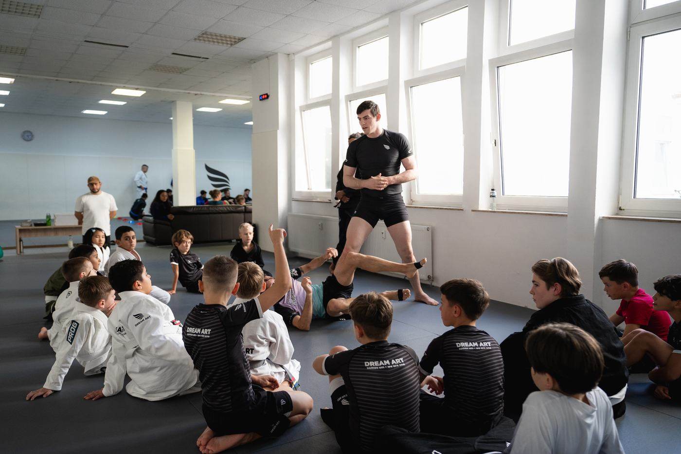 Die Kinderklasse sitzt im Kreis auf der Matte und hört dem Trainer zu